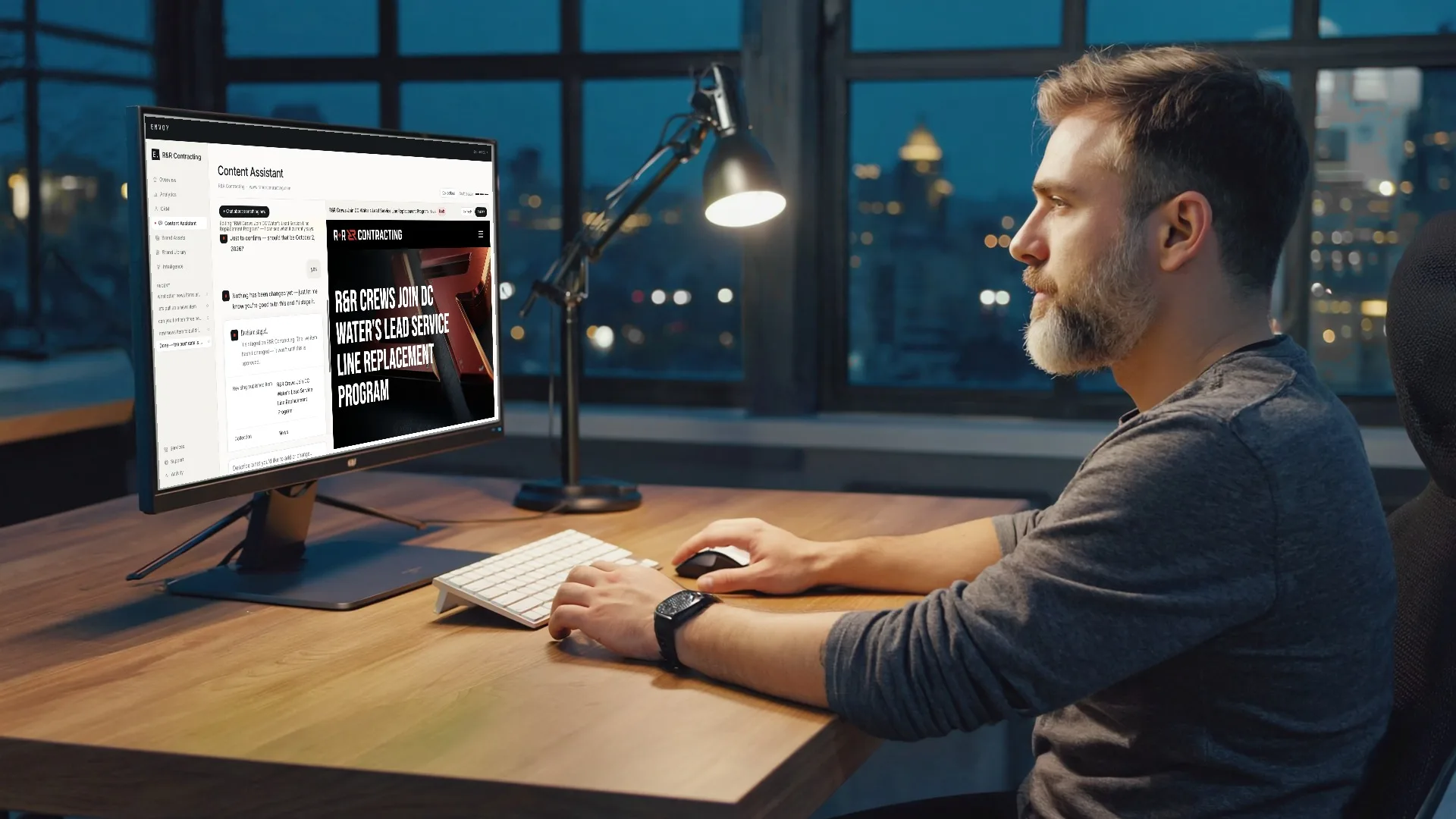 A marketing executive at his desk at dusk, working in the Envoy content assistant on a large monitor.